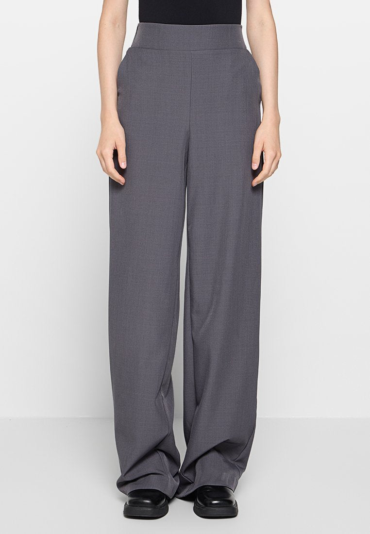Graue weitgeschnittene Hosen mit glatter Textur, hoher Taille und minimalistischem Design. Sie zeichnen sich durch klare Linien und dezente Drapierung aus und werden mit schwarzen Schuhen kombiniert.