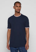 Mann in einem marineblauen T-Shirt mit hellrosa und weißem Besatz an Ärmeln und Kragen, steht vor einem schlichten hellgrauen Hintergrund.