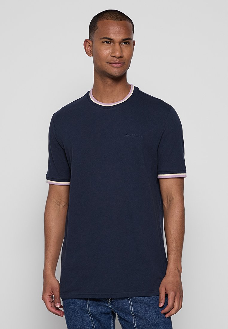 Mann in einem marineblauen T-Shirt mit hellrosa und weißem Besatz an Ärmeln und Kragen, steht vor einem schlichten hellgrauen Hintergrund.