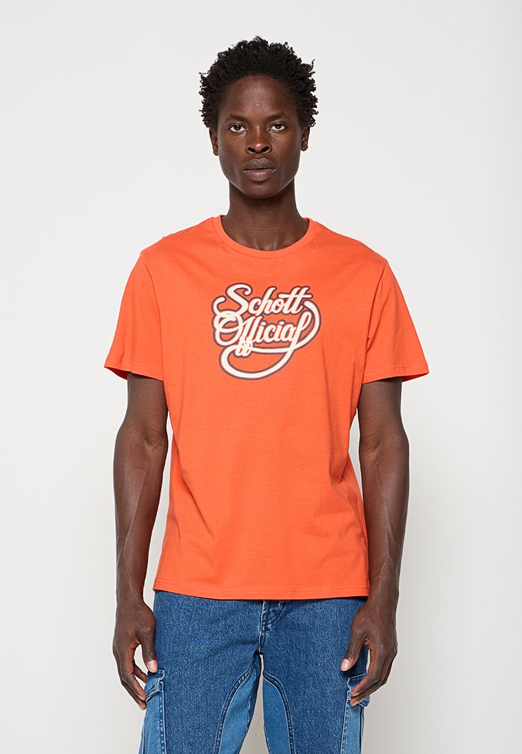 Mann trägt ein oranges T-Shirt mit weißem "Schott Official"-Schriftzug und blaue Jeans aus Denim und steht vor einem einfachen hellen Hintergrund.