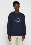 Junger Mann in einem dunkelblauen Sweatshirt mit glänzendem "DIESEL"-Schriftzug und hellbeigen, getragenen Jeans, der vor einem schlichten, hellen Hintergrund steht.