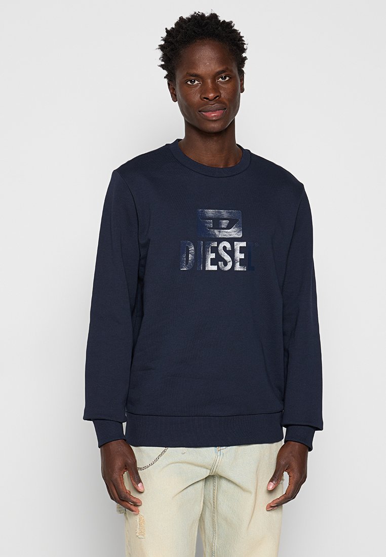 Junger Mann in einem dunkelblauen Sweatshirt mit glänzendem "DIESEL"-Schriftzug und hellbeigen, getragenen Jeans, der vor einem schlichten, hellen Hintergrund steht.