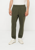 Mann trägt olivgrüne Jogginghosen mit elastischen Bündchen und weißen Sneakers auf einem einfarbigen weißen Hintergrund.