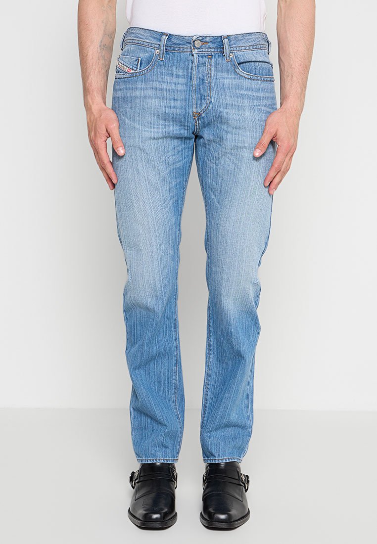 Mann trägt hellblaue Jeans mit geradem Bein, weißes Hemd und schwarze Lederschuhe, steht vor einem einfarbigen Hintergrund.