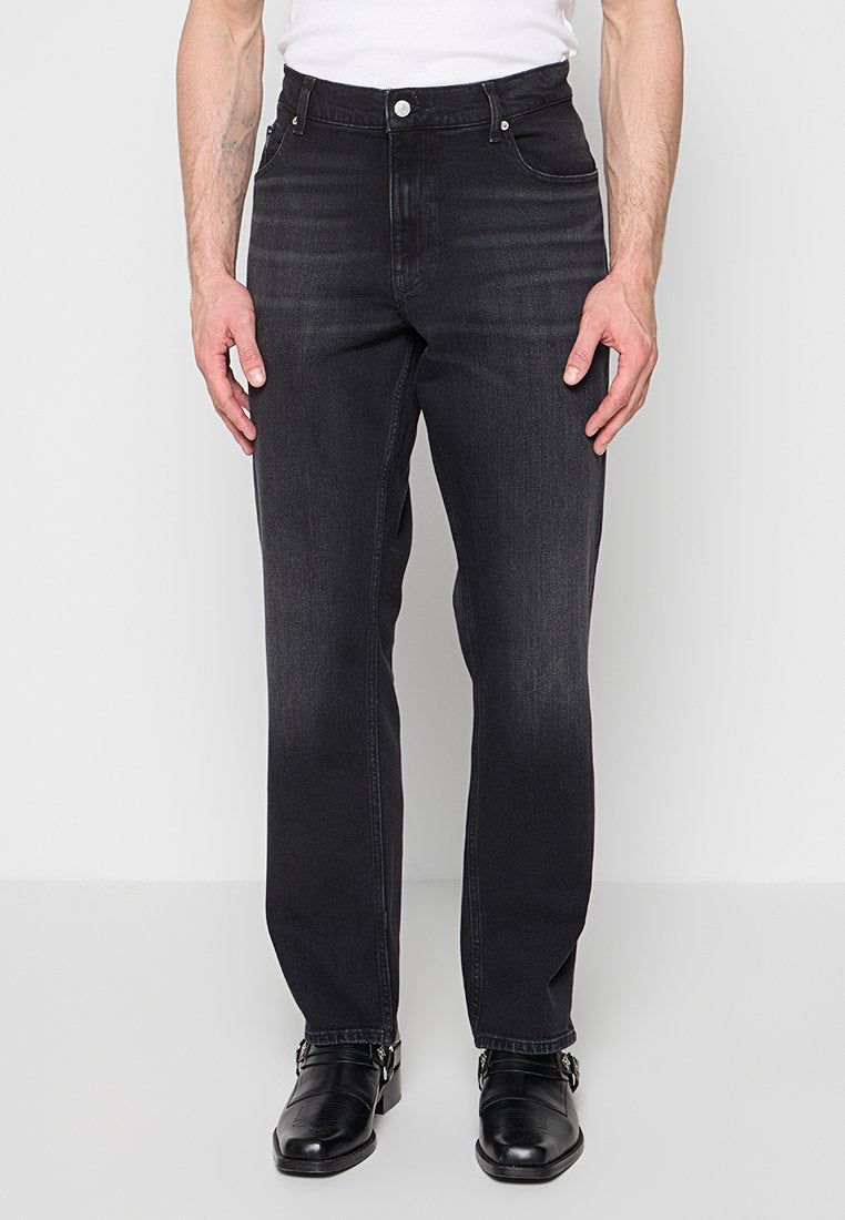 Mann trägt ausgewaschene schwarze Jeans mit geraden Beinen, weißes Hemd und schwarze Lederschuhe, steht vor einem schlichten hellen Hintergrund.