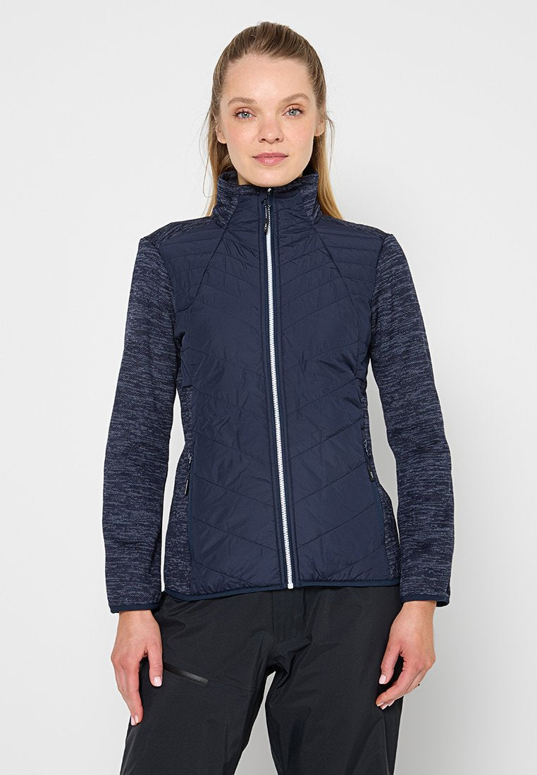Frau trägt eine marineblaue gesteppte Jacke mit gemusterten Ärmeln und schwarze Hosen, steht vor einem schlichten hellen Hintergrund.