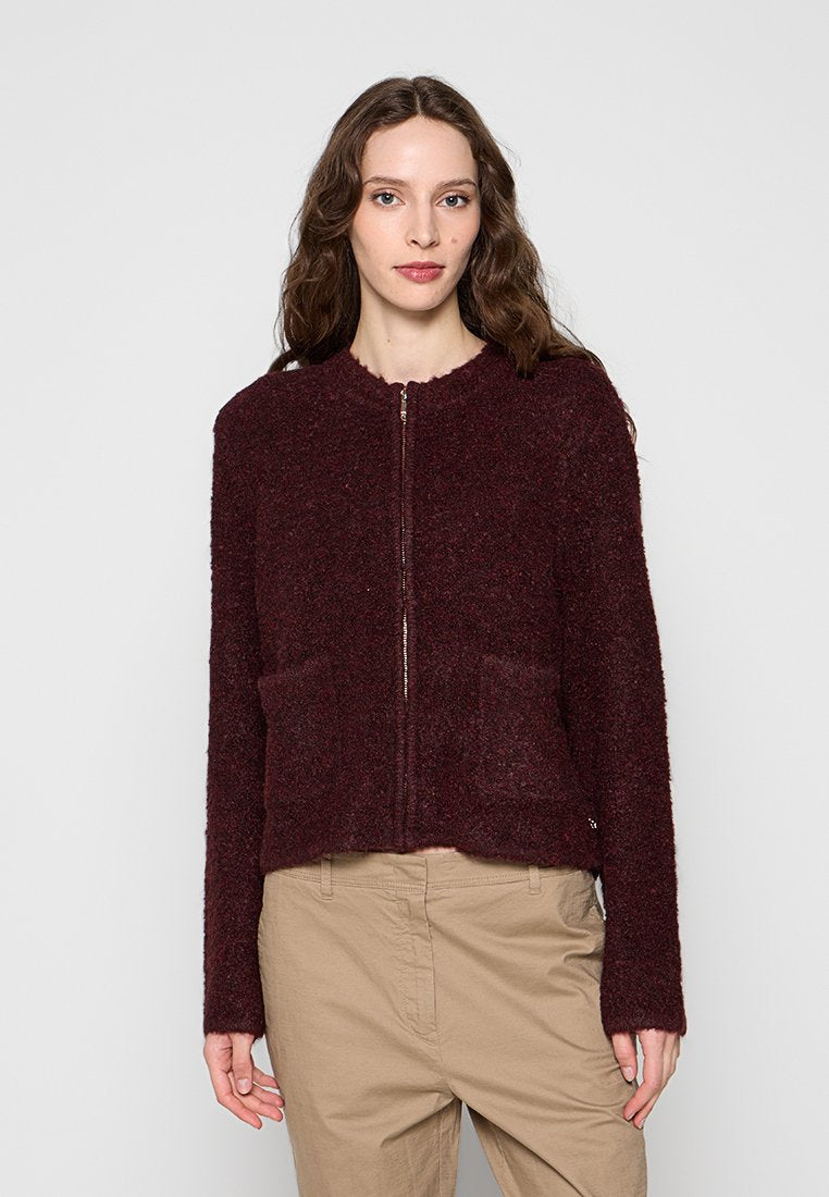 Junge Frau mit langen braunen Haaren, die eine dunkelburgunderfarbene, strukturierte Jacke mit Reißverschluss und beige Hosen vor einem einfarbigen Hintergrund trägt.