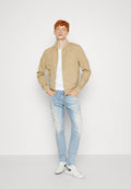 Beige Bomberjacke mit einer glatten Oberfläche, weißes T-Shirt, hellblaue Jeans und weiße Sneaker mit schwarzen Akzenten. Stehende Pose vor einem schlichten Hintergrund.