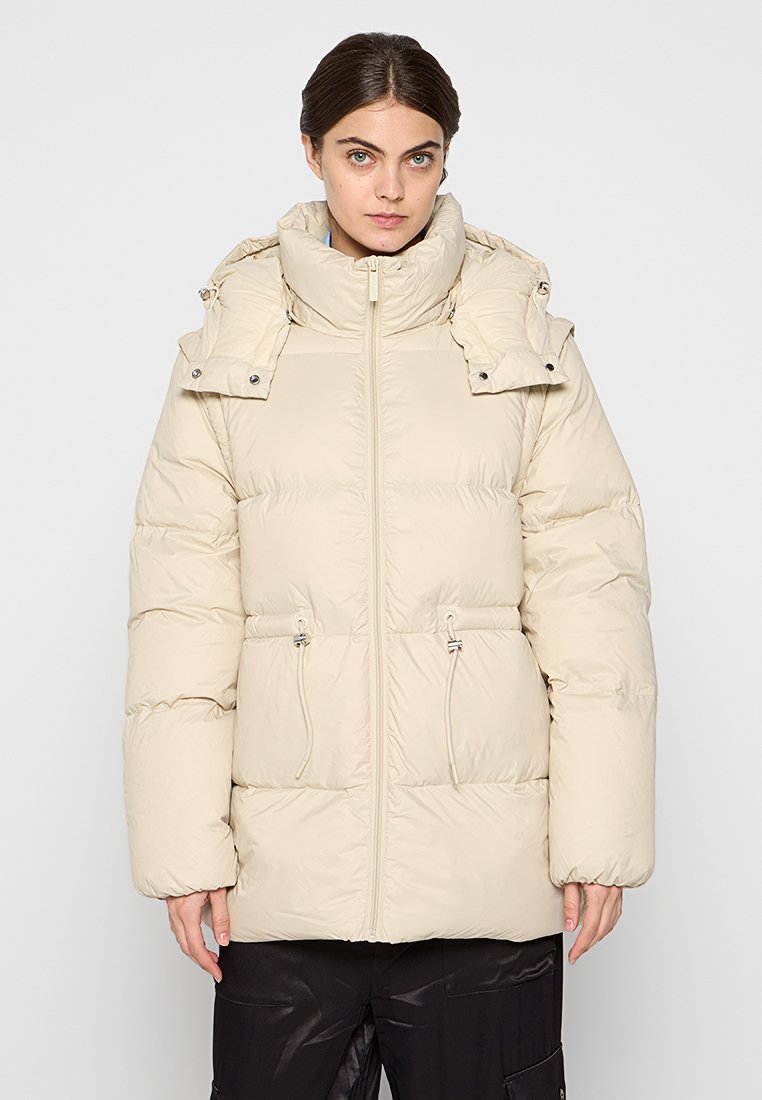 Beige Steppjacke mit hohem Kragen, Reißverschluss, verstellbaren Kordeln in der Taille und Druckknopfakzenten an der Kapuze.