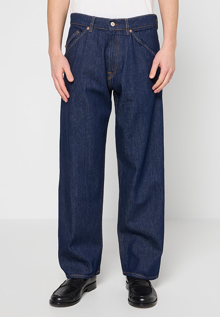 Locker sitzende dunkelblaue Jeans aus Denim, getragen mit schwarzen Lederschuhen und einem weißen Hemd, von der Mitte des Oberkörpers bis zu den Füßen zugeschnitten.