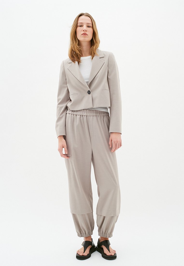 Junge Frau steht vor einem weißen Hintergrund und trägt ein beiges, cropped Blazer, passende elastische Hosen, ein weißes Shirt und schwarze Sandalen mit offenen Zehen.