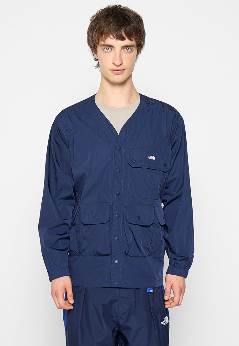 Navyblaue, leichte Jacke mit V-Ausschnitt, langen Ärmeln, zwei Fronttaschen und Logo auf der Brust. Glatte Textur und Druckknopfverschluss.