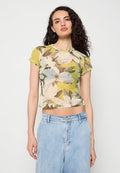 Junge Frau mit lockigem Haar, die ein gelbes, florales transparentes Crop-Top und hellblaue High-Waist-Jeans vor neutralem Hintergrund trägt.