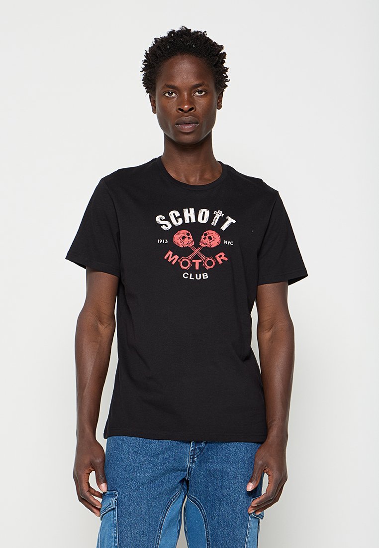 Junger Mann trägt ein schwarzes T-Shirt mit Totenkopf-Grafik und blauen Jeans, steht vor einfachem weißem Hintergrund.