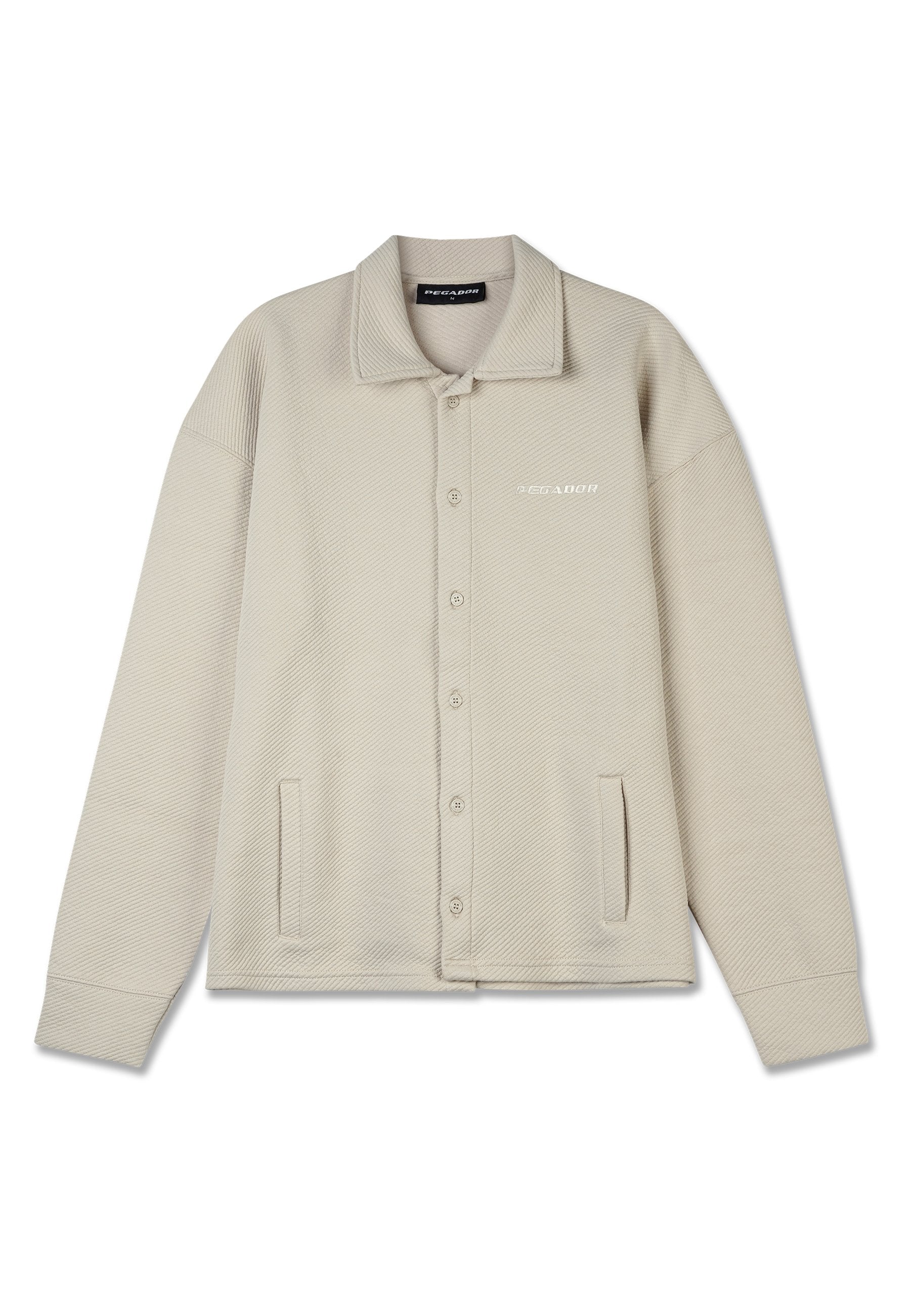 Beige Hemdjacke mit strukturiertem Muster, großem Kragen, zwei Fronttaschen und einem dezenten Logo auf der Brust. Weicher Stoff.