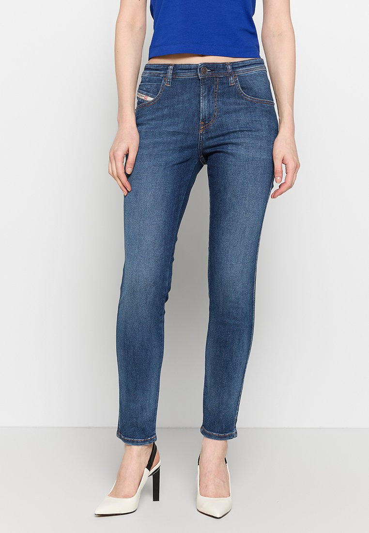 Dunkelblaue Skinny Jeans aus Denim, mit frontalen Taschen und orangenen Nähten. Getragen mit einem blauen Oberteil und weißen High Heels.
