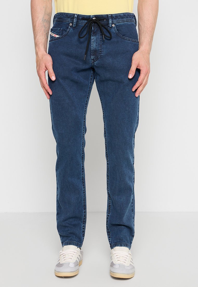 Mann trägt blaue Jeans mit schwarzem Kordelzug und weißen Sneakers, steht vor einem schlichten weißen Hintergrund.