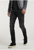 Mann in schwarzen, eng anliegenden Jeans, schwarzen Sneakers mit weißen Sohlen, schwarzem T-Shirt und grauer Jeansjacke, stehend vor einem einfarbigen Hintergrund.