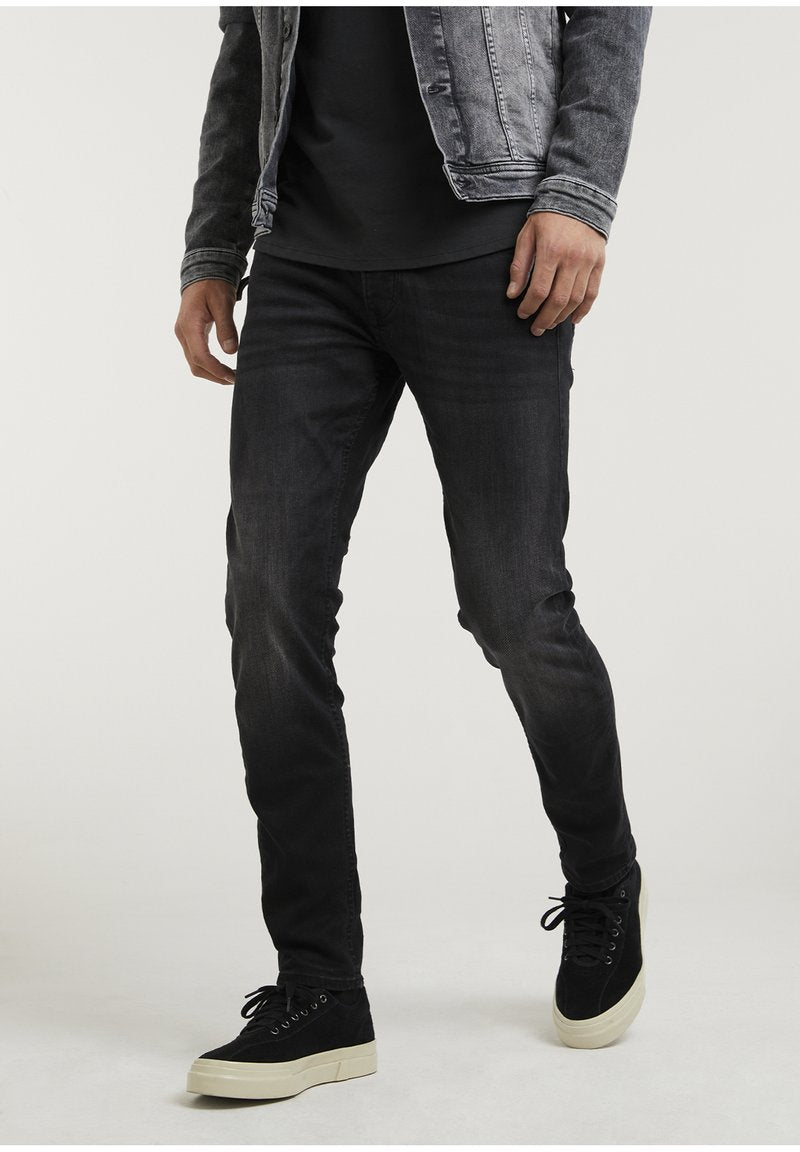 Mann in schwarzen, eng anliegenden Jeans, schwarzen Sneakers mit weißen Sohlen, schwarzem T-Shirt und grauer Jeansjacke, stehend vor einem einfarbigen Hintergrund.