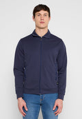 Mann trägt eine marineblaue Jacke mit Reißverschluss und Kragen sowie eine blaue Jeans und steht vor einem schlichten hellgrauen Hintergrund.