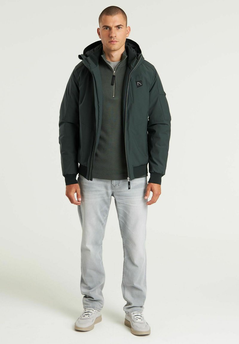Dunkelgrüne Zip-Jacke mit Kapuze, ausgestattet mit einem strukturierten Design, Rippbündchen und einem kleinen Logo auf der Brust, kombiniert mit hellgrauen Jeans.