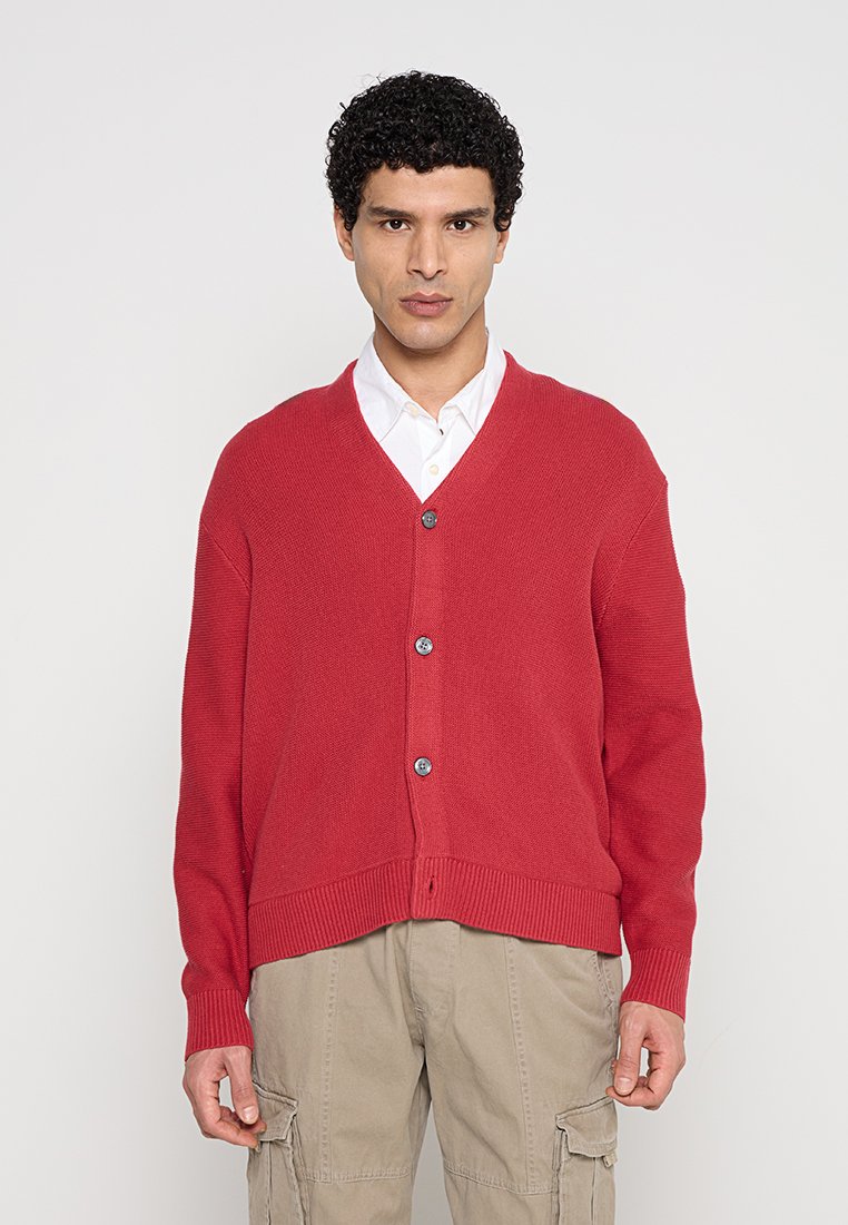 Mann trägt einen roten Knopfstrickpullover über einem weißen Hemd und beige Cargo-Hosen, steht vor einem schlichten hellen Hintergrund.