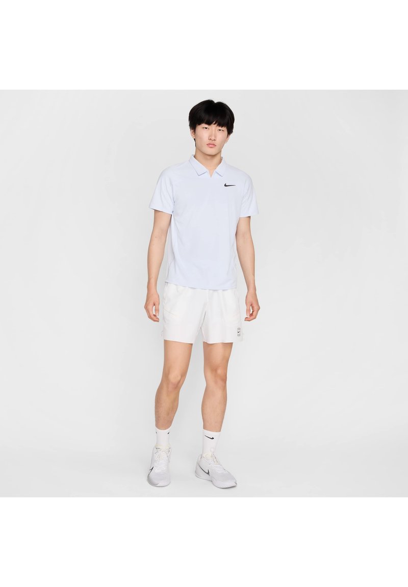 Helles blaues Polohemd mit kurzen Ärmeln und schwarzem Nike-Logo, kombiniert mit weißen Shorts. Das Modell trägt weiße Schuhe und Socken.