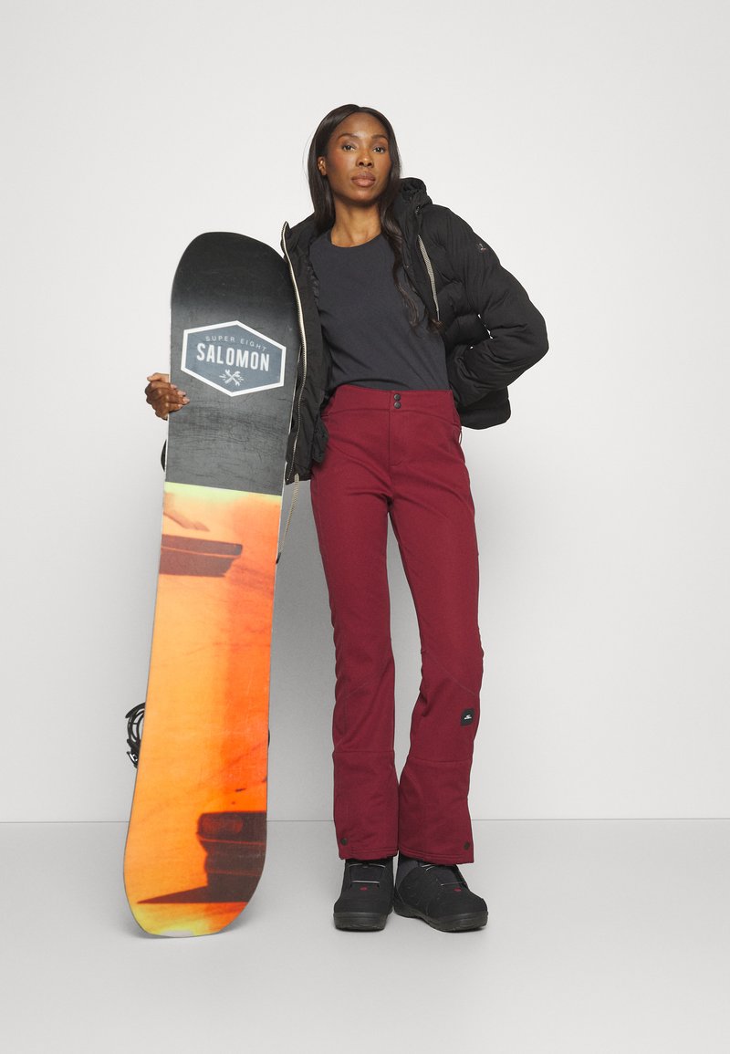 Frau in schwarzer Jacke, dunkler Bluse und roten Schneehosen, die ein Snowboard mit schwarzem und orangefarbenem Design hält, steht vor einem weißen Hintergrund.