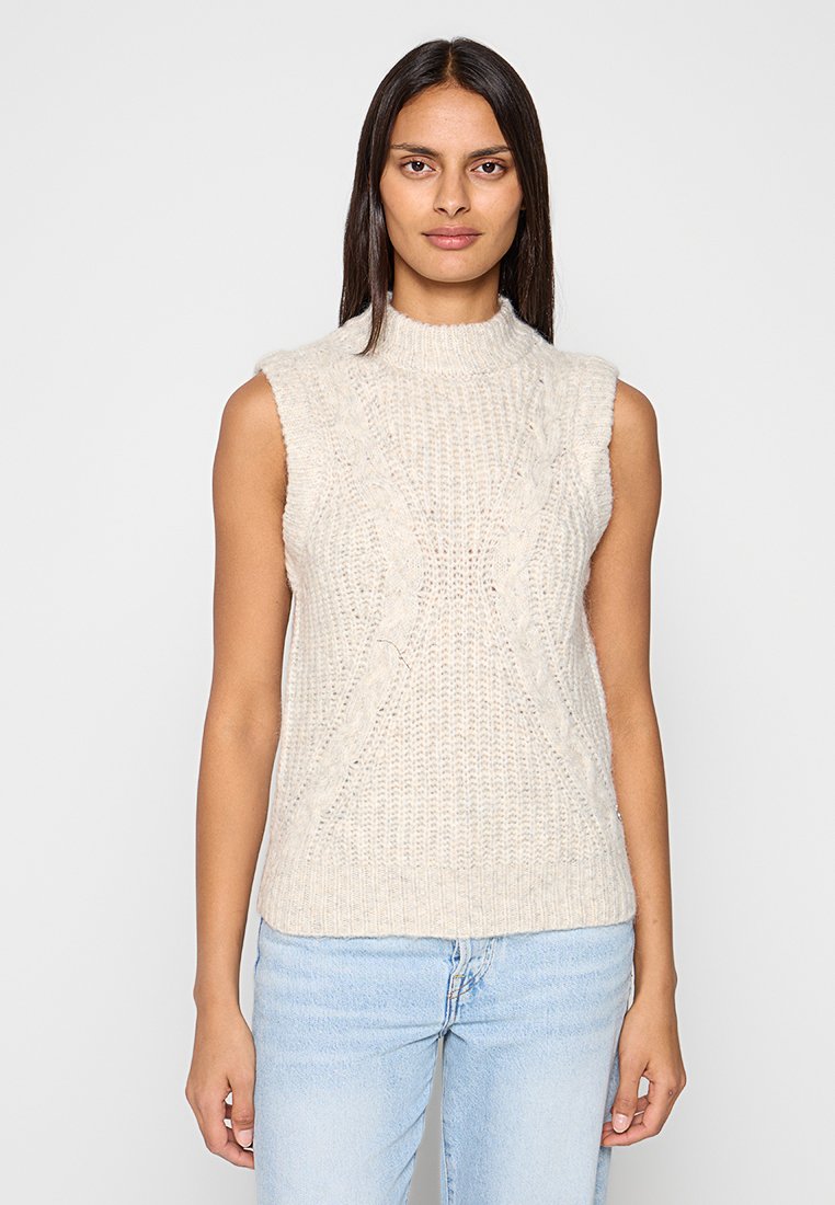 Ärmelloses Strickoberteil in Creme mit hohem Kragen, strukturierten Zopfmustern und figurbetontem Design. Kombiniert mit hellblauen Jeans.