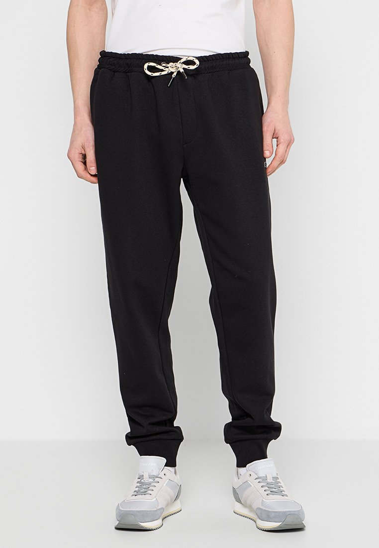 Schwarze Jogginghosen mit elastischem Bund, Kordelzug und schmal zulaufenden Säumen. Getragen mit grauen und weißen Sportschuhen.