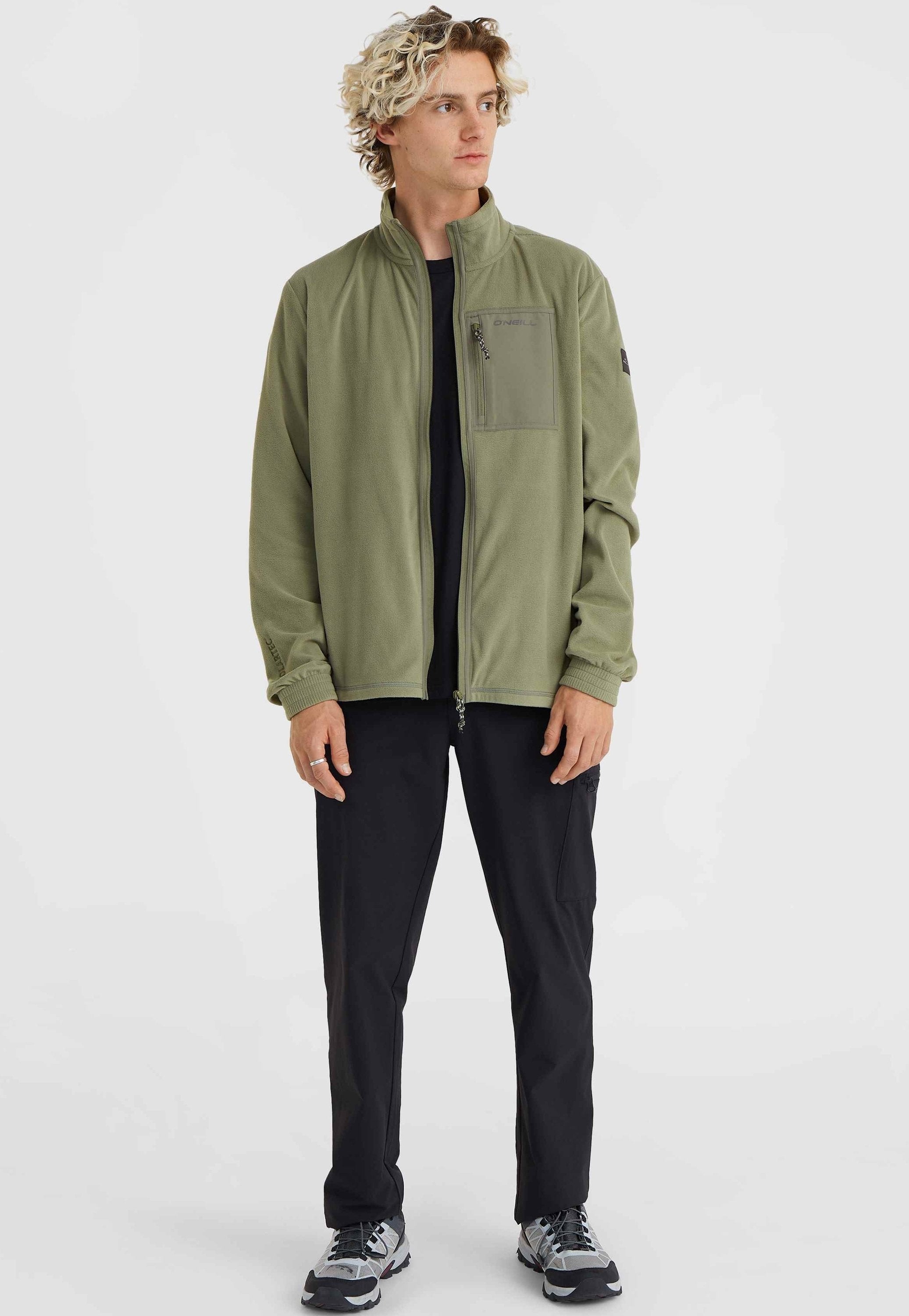 Grüne Fleecejacke mit hohem Kragen, Frontreißverschluss und Brusttasche. Schwarze Hose und graue Sportschuhe vervollständigen das Outfit.