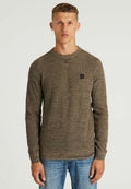 Braun strukturierter Pullover mit Rundhalsausschnitt, gerippten Bündchen und einem kleinen schwarzen Logo-Patch auf der Brust, kombiniert mit blauen Jeans.