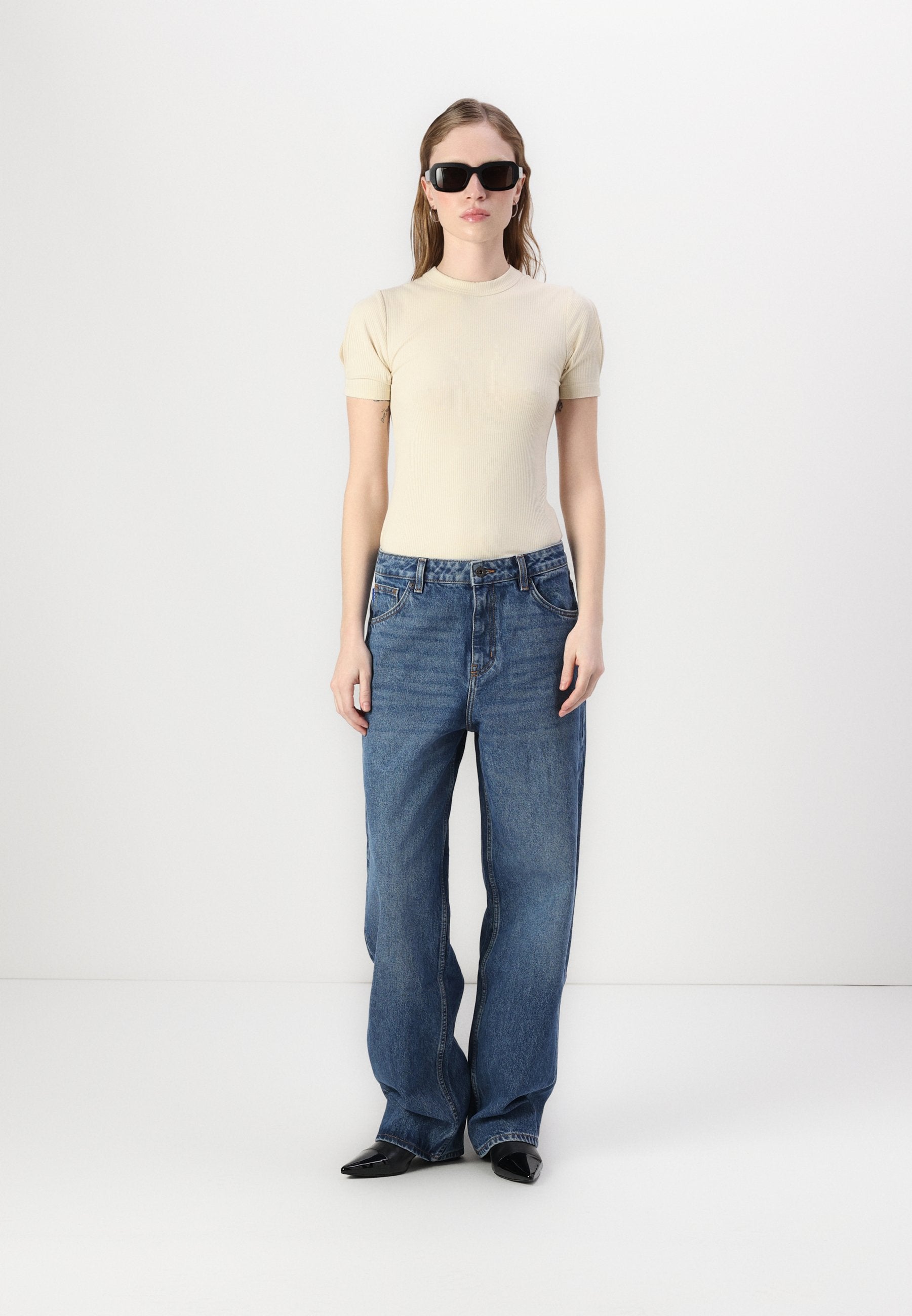 Beiges, geripptes Kurzarmoberteil kombiniert mit blauen Jeans mit weitem Bein. Accessoires: schwarze Sonnenbrille und schwarze spitze Schuhe.