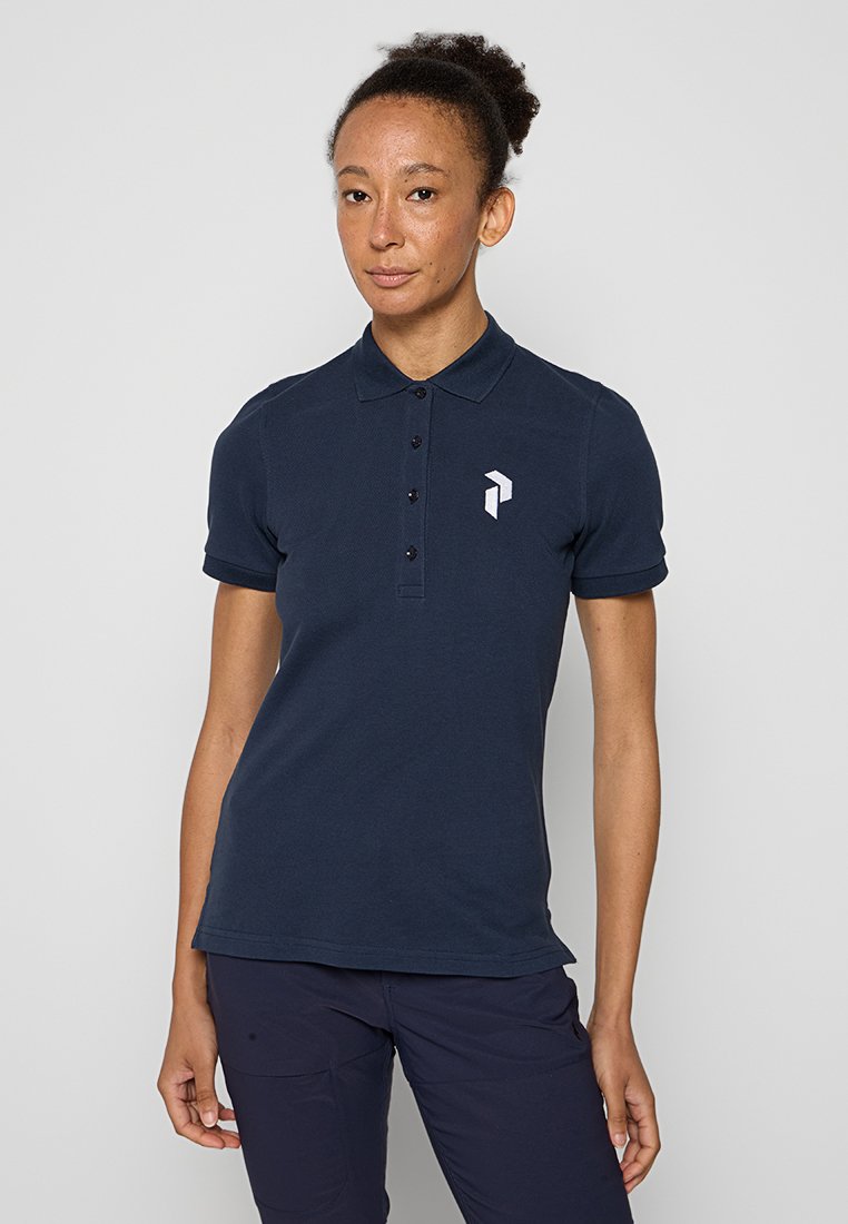 Frau mit lockigem Haar in einem marineblauen Poloshirt mit einem weißen geometrischen Logo auf der Brust, vor einem einfarbigen hellen Hintergrund stehend.