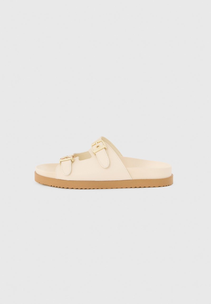 Beige Slidesandale mit zwei verstellbaren goldenen Schnallenriemen und einer flachen, braunen Gummisohle auf hellgrauem Hintergrund.
