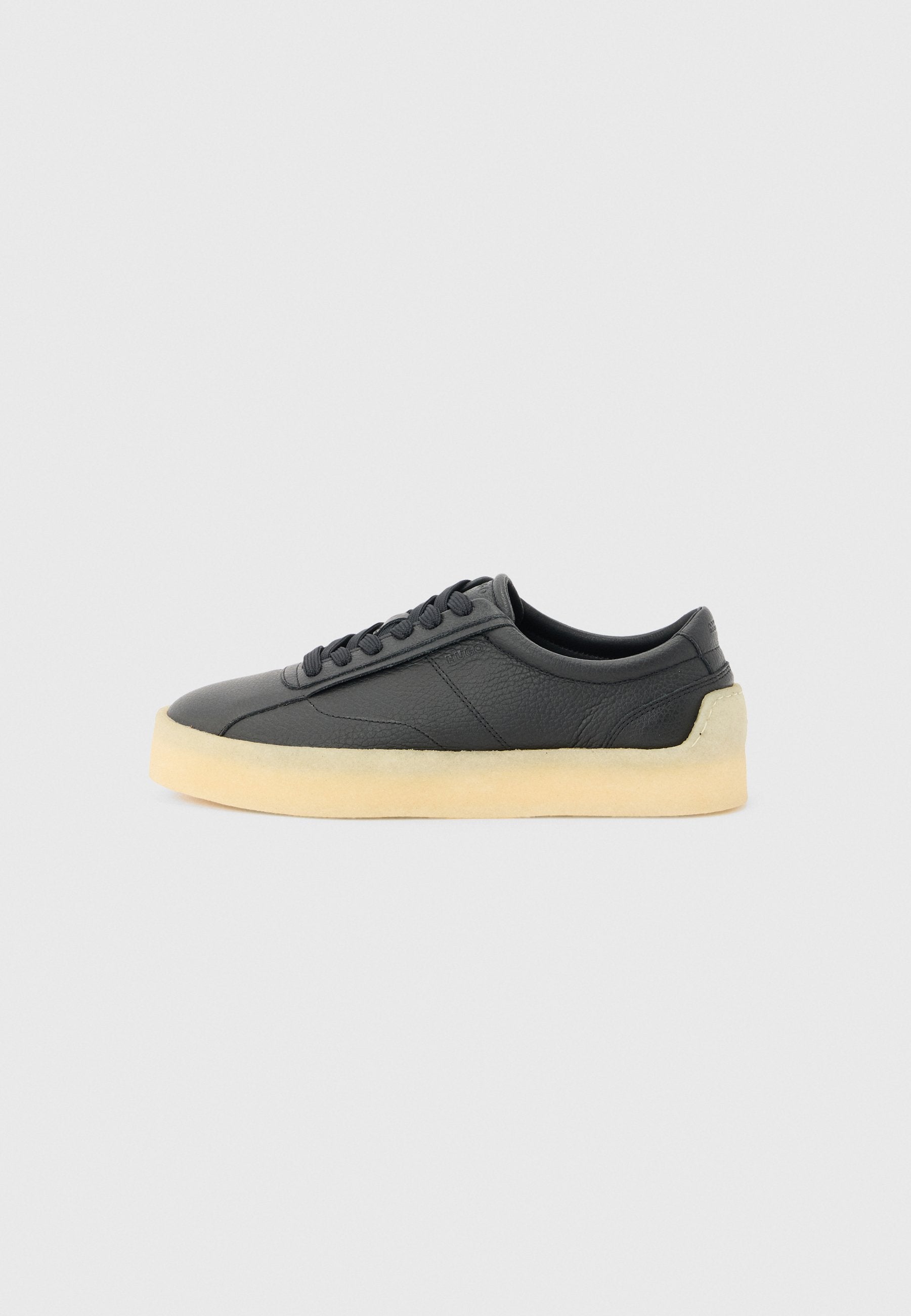 Schwarzer Low-Top-Ledersneaker mit abgerundeter Spitze, schwarzen Schnürsenkeln und dicker, cremeweißer Gummisohle, seitlich auf hellem grauen Hintergrund dargestellt.