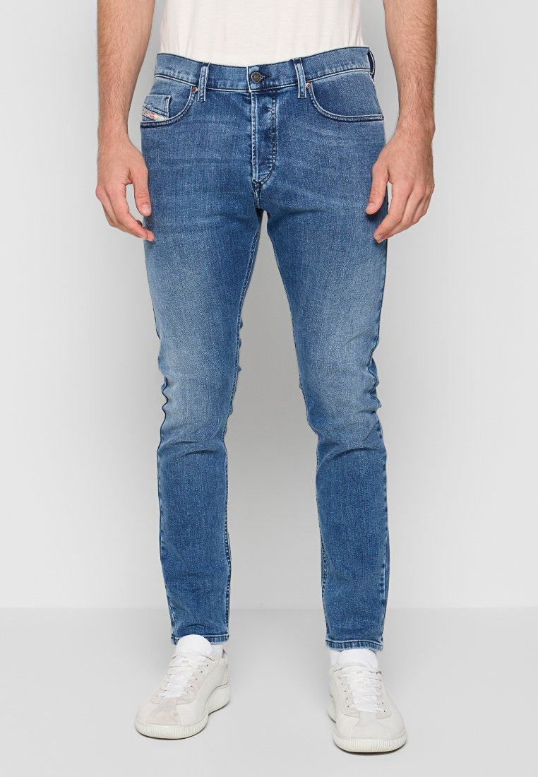 Mann in schlank geschnittenen blauen Jeans mit leichter Ausbleichung, weißen Socken und weißen Sneakers, vor einem schlichten hellgrauen Hintergrund stehend.