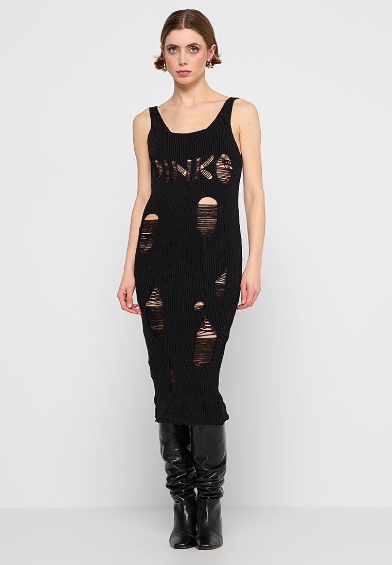 Frau in einem ärmellosen schwarzen Strickkleid mit Aussparungen und "PINKO"-Schriftzug, kombiniert mit schwarzen hohen Lederstiefeln, die vor einer weißen Wand steht.