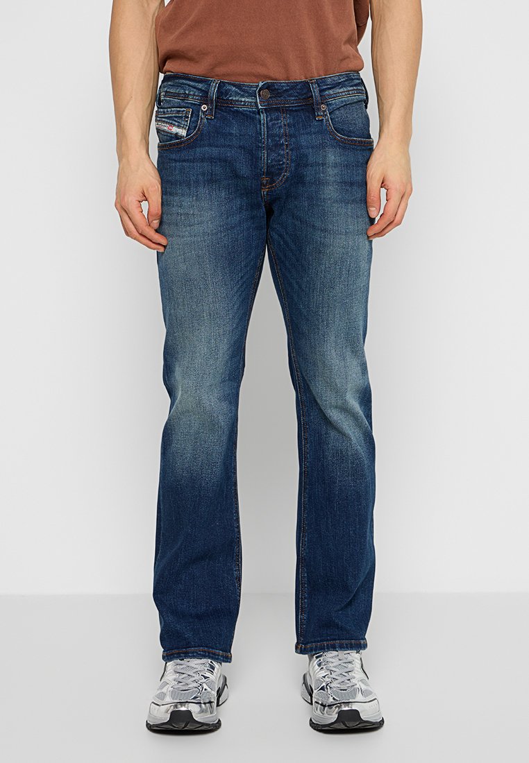 Person in geraden, ausgebleichten blauen Jeans und weiß-grauen Sportschuhen, der vor einem schlichten hellen Hintergrund steht.