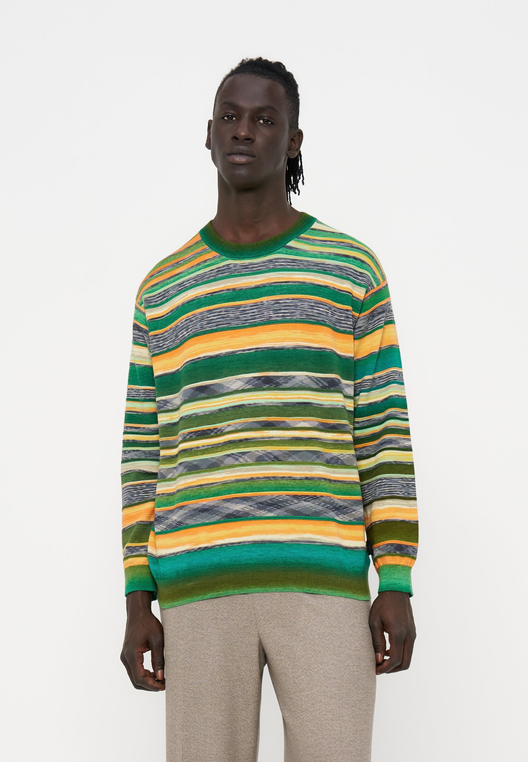 Mann mit dunkler Haut und Dreadlocks trägt einen bunten gestreiften Pullover mit grünen, gelben und orangen Tönen und beigen Hosen und steht vor weißem Hintergrund.