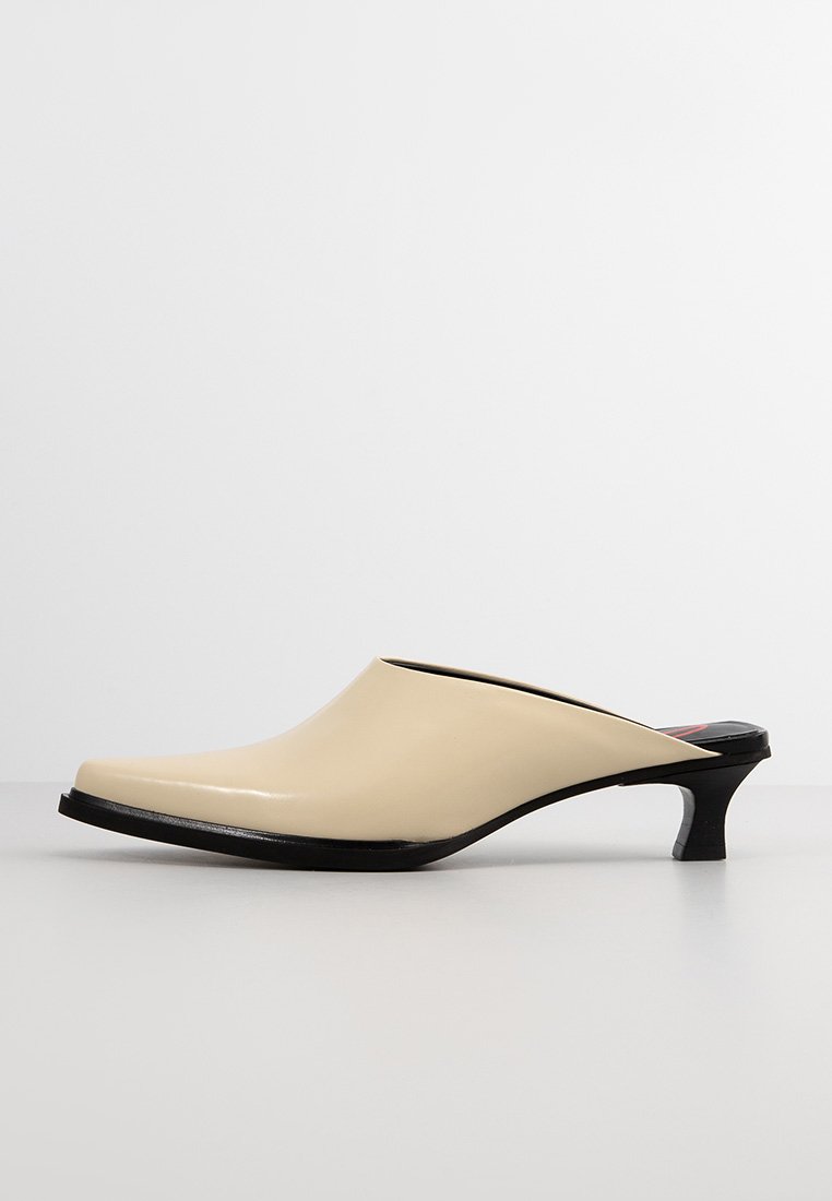Beige glänzende Mules mit einer spitzen Zehenform, schlankem Design, niedrigem schwarzen Absatz und offener Rückseite. Glatte Textur ohne sichtbare Muster oder Akzente.