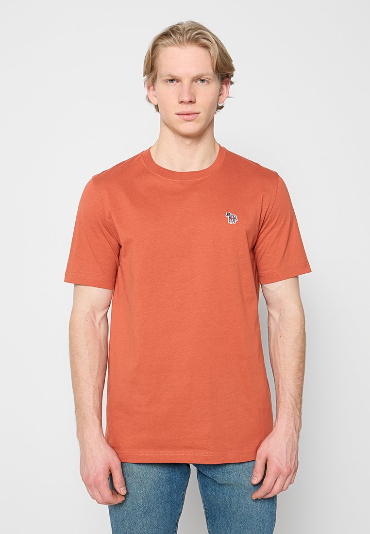 Baumwoll-T-Shirt in Rostorange mit kurzen Ärmeln, rundem Ausschnitt und einem kleinen blau-weißen Logo auf der linken Seite. Getragen zu blauen Jeans.