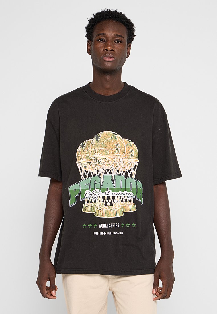 Ein junger Mann steht mit dem Gesicht zur Kamera und trägt ein schwarzes, übergroßes T-Shirt mit einer Basketball-Trophäe und dem Text "Pecador College Association".