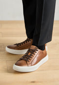 Braune Ledersneaker mit einer weißen Gummisohle, ausgestattet mit braunen Schnürsenkeln und einem Logodetail. Getragen mit schwarzen Hosen und einer schwarzen Socke.