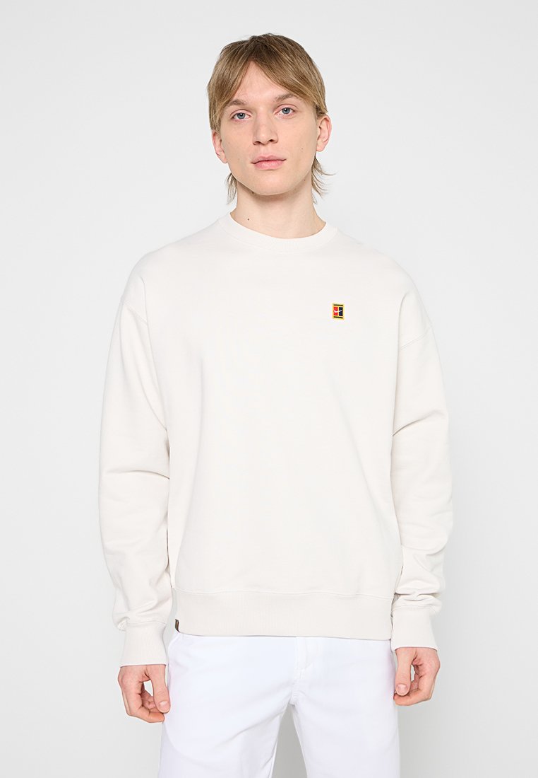 Junger Mann mit hellem Haar, der einen schlichten weißen Pullover mit einem kleinen bunten Logo auf der linken Brust und weiße Hosen trägt.