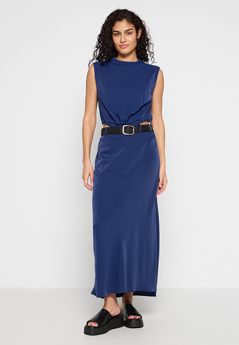 Schickes marineblaues Maxikleid mit hohem Ausschnitt, gerafftem Design an der Taille und einem schwarzen Gürtel. Kombiniert mit schwarzen Plateauschuhen zum Hineinschlüpfen.