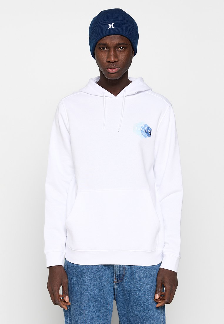 Junger Mann trägt einen weißen Hoodie mit blauem Logo und eine dunkelblaue Mütze und steht vor einem einfarbigen hellen Hintergrund.