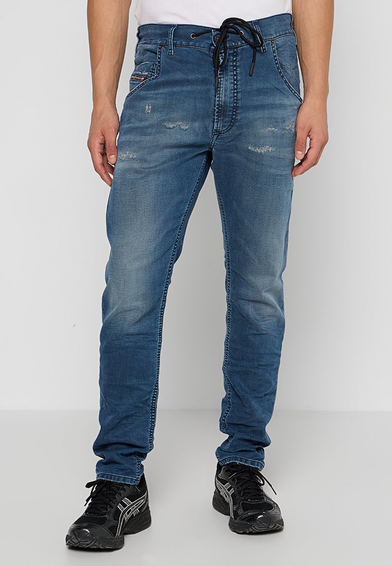 Blaue Denim-Jeans mit einer lockeren Passform, mit einem Kordelzug im Bund, distressed Details und sichtbaren Nähakzenten. Kombiniert mit schwarzen Sneakers.