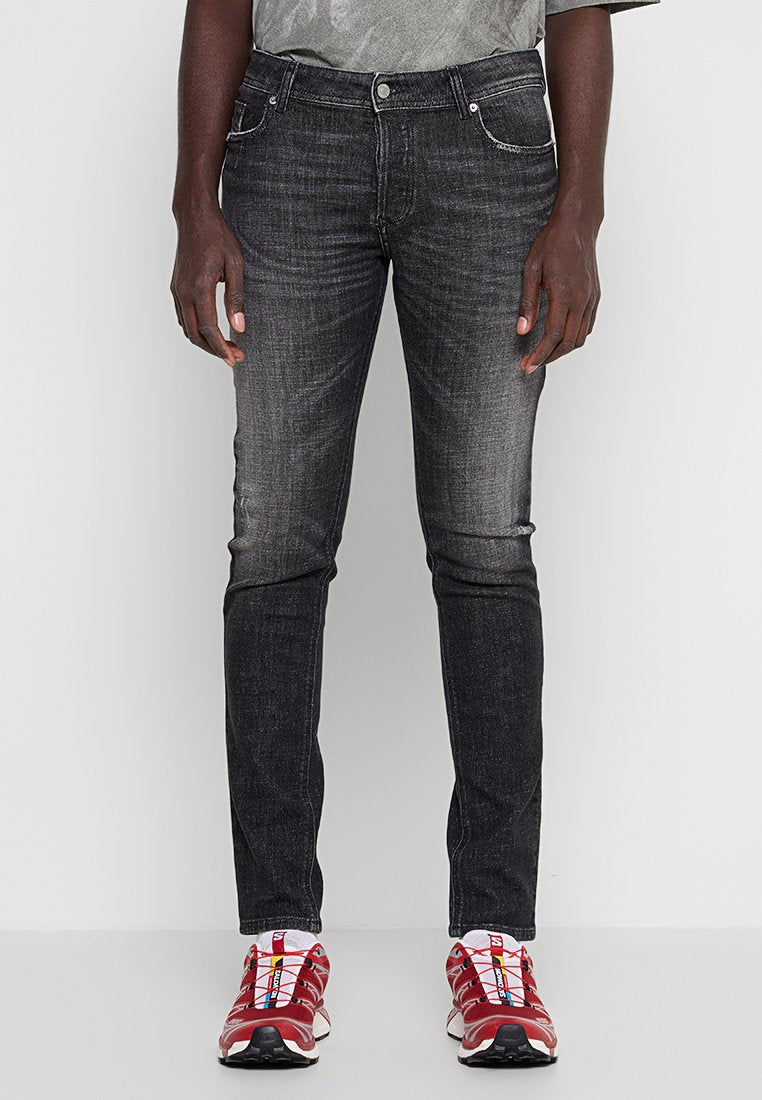 Mann trägt ausgewaschene schwarze Slim-Fit-Jeans und rote Sportschuhe, steht vor einem einfarbigen hellen Hintergrund.
