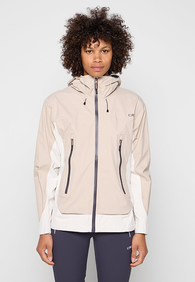Frau mit lockigem Haar, die eine beige-weiße Kapuzenjacke mit Reißverschluss und dunkle Leggings trägt, steht vor einem schlichten Hintergrund.
